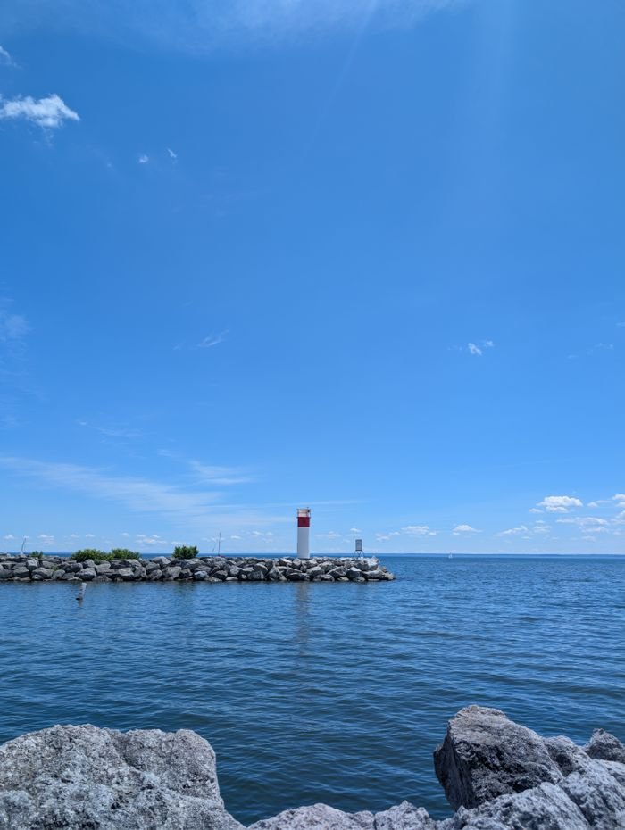 A red and white lighthouse standing on a rocky pier under a bright blue sky with ocean waves.